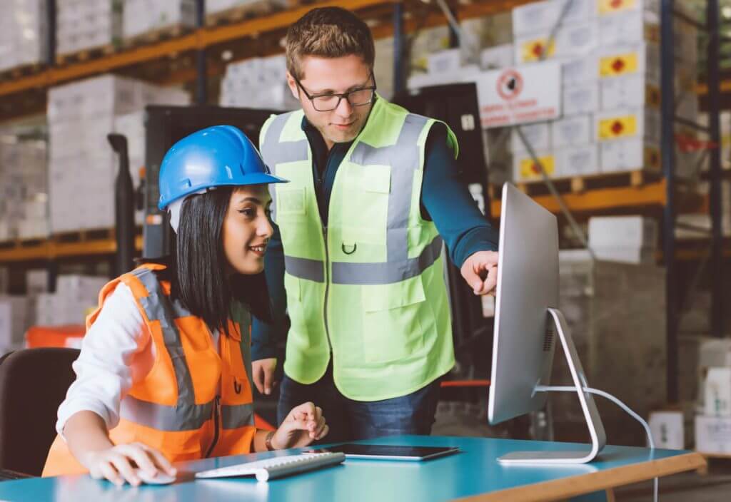 Two people wearing helmets and reflective vests looking at a computer